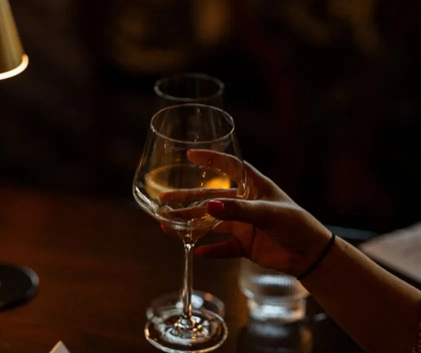 Manicured hand with red nails holds amber wine in dim light.