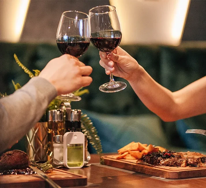 Close-up of hands toasting red wine over steak and potatoes.