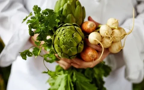Chef's hands holding fresh artichokes, cilantro, shallots, and small white turnips.