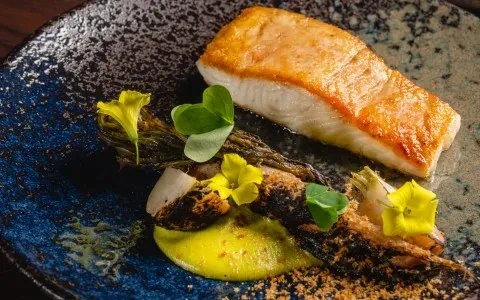 Seared fish, green vegetables, yellow sauce, and edible flowers on a plate.