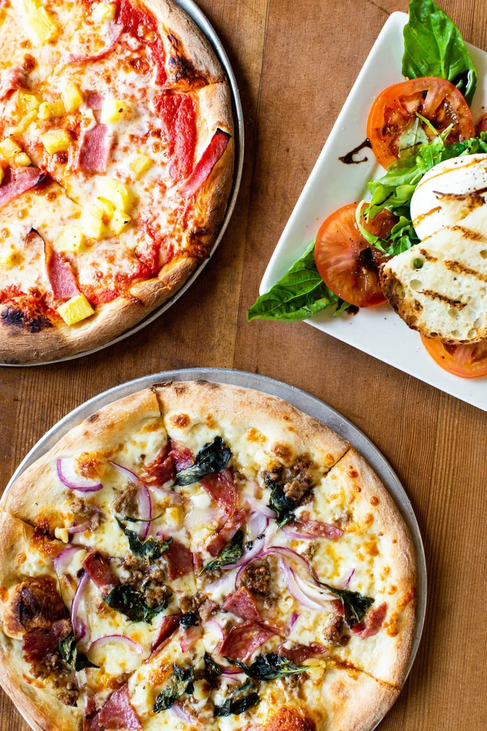 Two pizzas, caprese salad, grilled bread on a wooden table.
