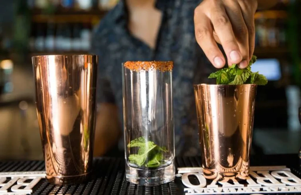 Bartender adds fresh mint to a copper shaker on a bar mat.