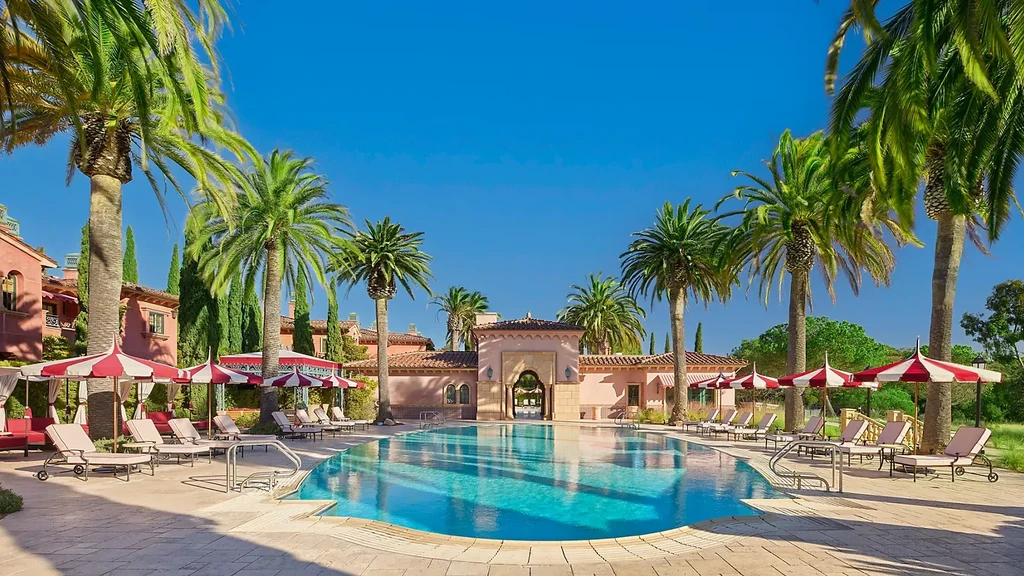 Empty resort pool with palm trees, lounge chairs, and terracotta buildings.