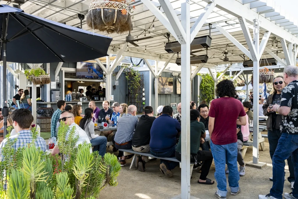 Crowded restaurant patio with pergola, bar, and sports on TV.