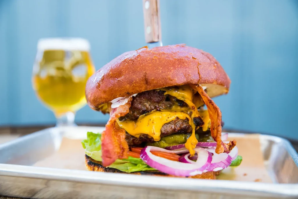 Detailed gourmet burger with cheese, bacon, vegetables, knife, and beer.