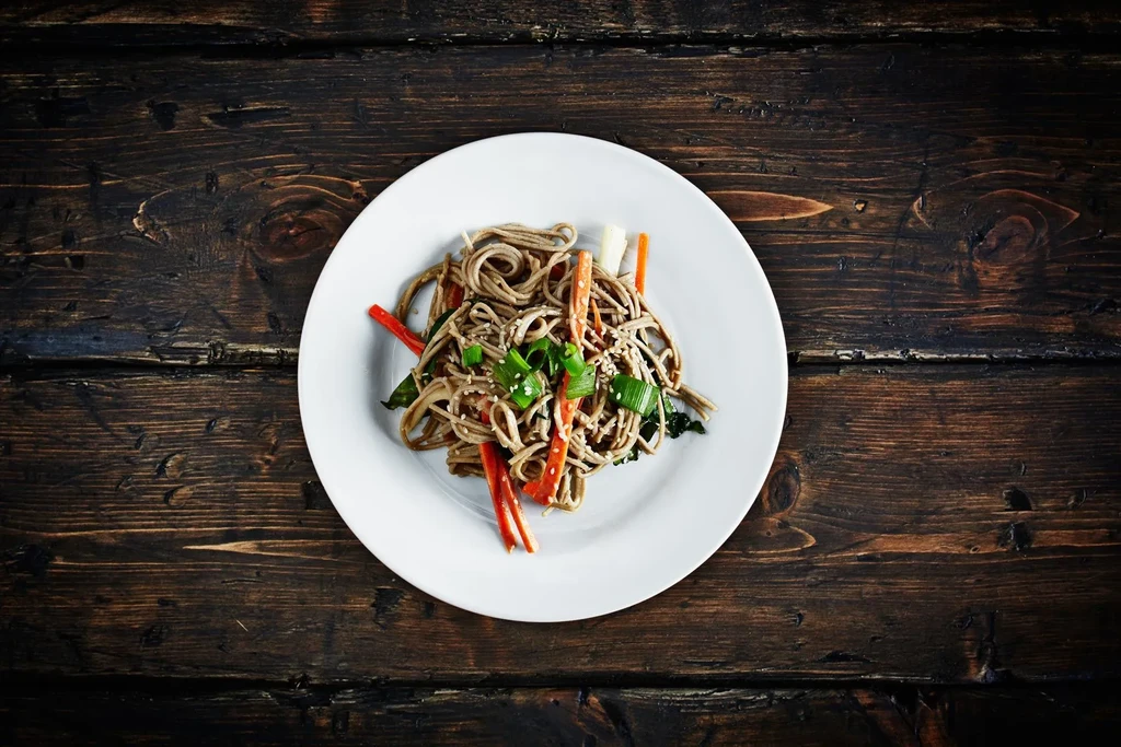 Overhead of buckwheat noodles with vegetables, sesame seeds on a wood table.