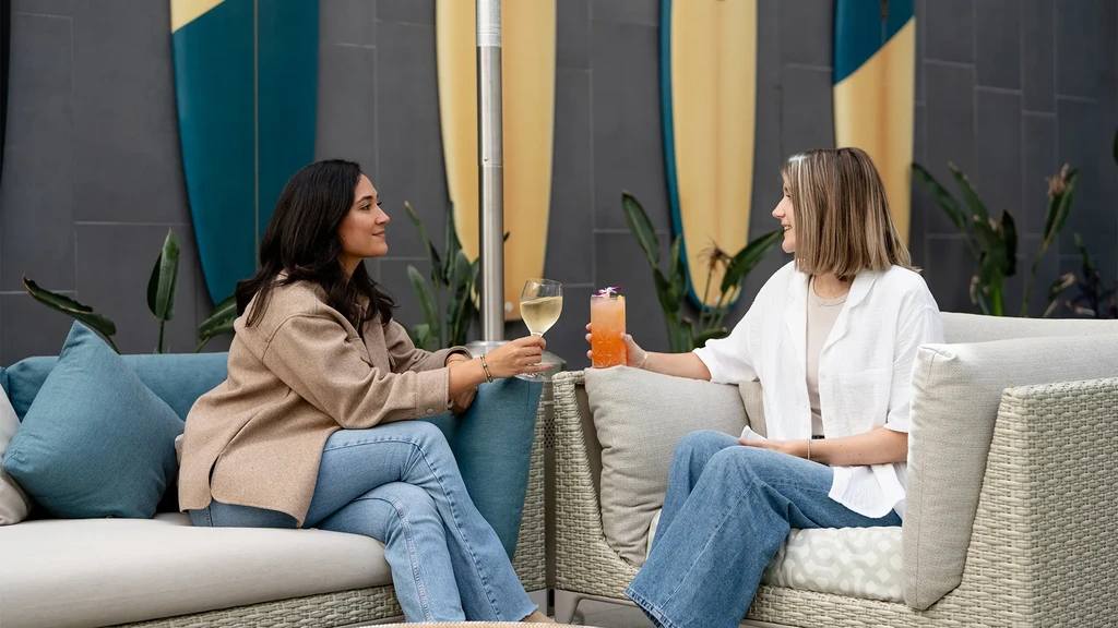 Two women converse, holding drinks, on outdoor sofas with surfboards.