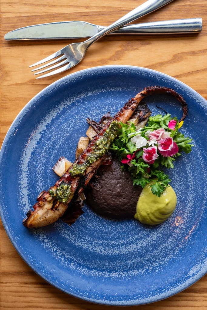 Grilled octopus, vegetables, green sauces, and flowers on a blue plate.