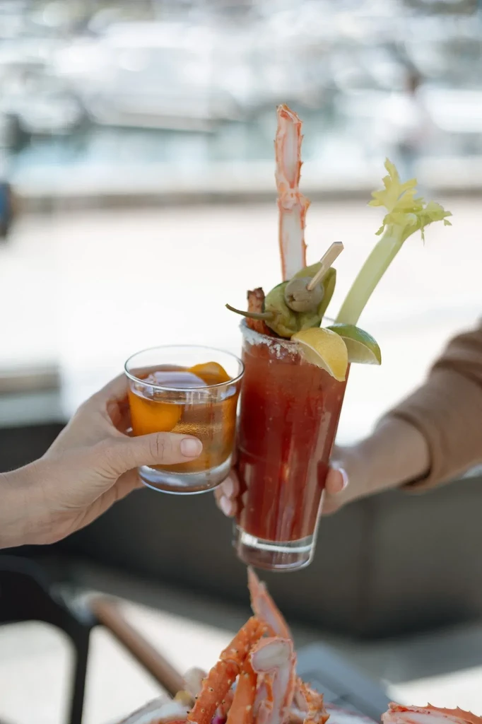 Hands clinking an amber cocktail and a heavily garnished Bloody Mary.