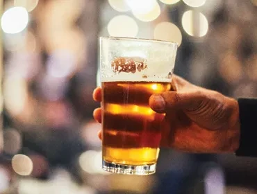 Hand holding glass of amber beer with frothy head, blurred lights.