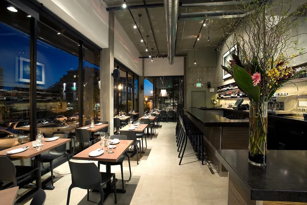 Modern restaurant interior with a long bar, dining tables, and floral arrangement.