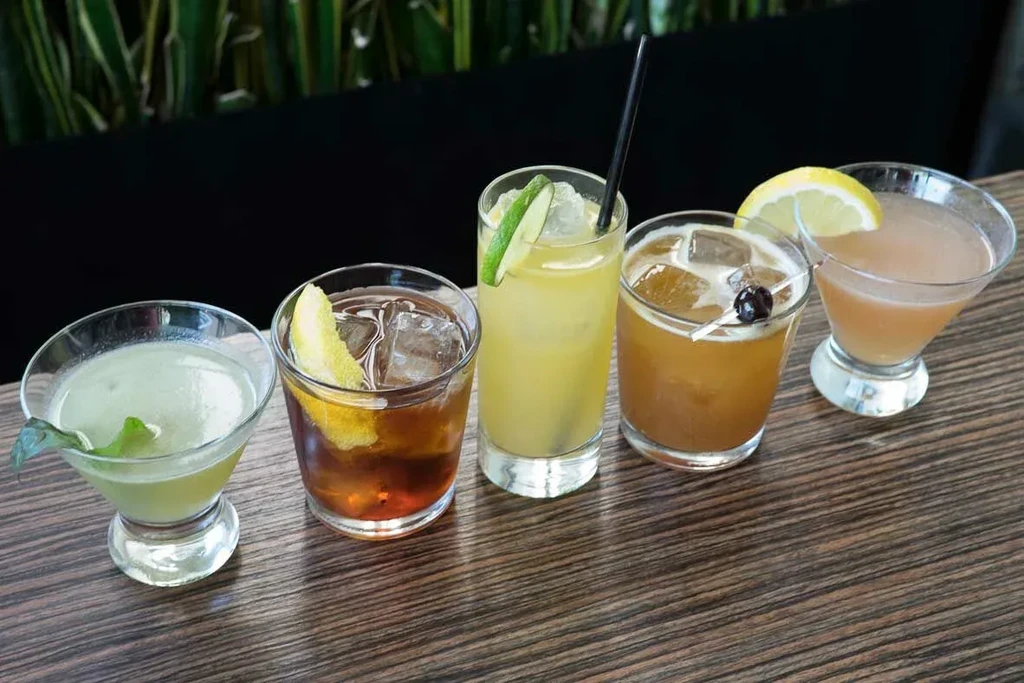 Five diverse cocktails with garnishes sit on a textured wooden table.