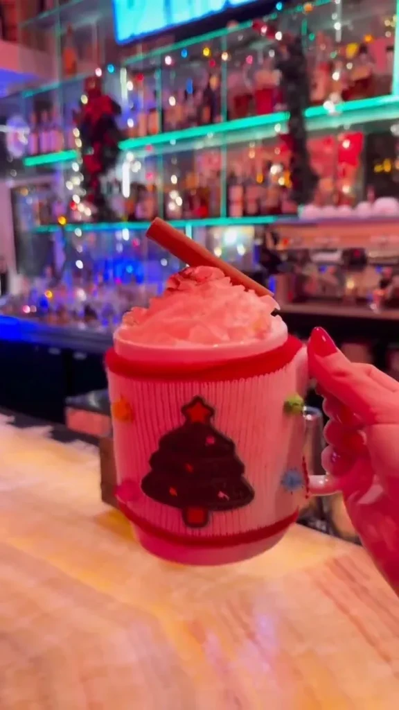 Hand holds a Christmas tree mug with whipped cream at a neon-lit bar.