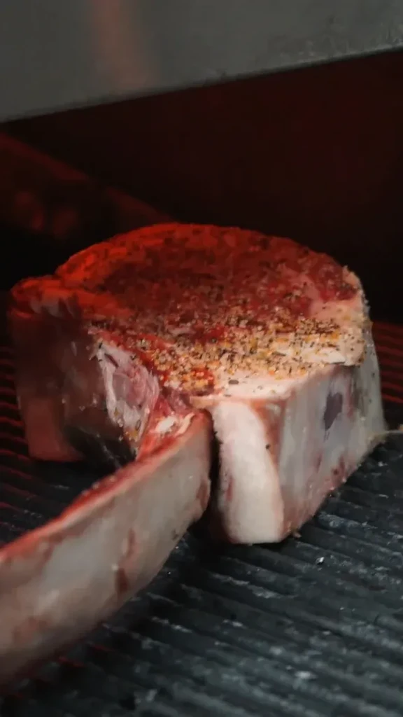 Raw, seasoned, bone-in steak on a grill grate under red light.