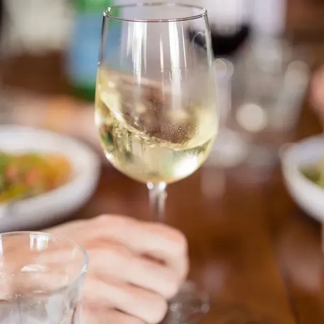 Hand holds wine glass with white wine at a dining table.