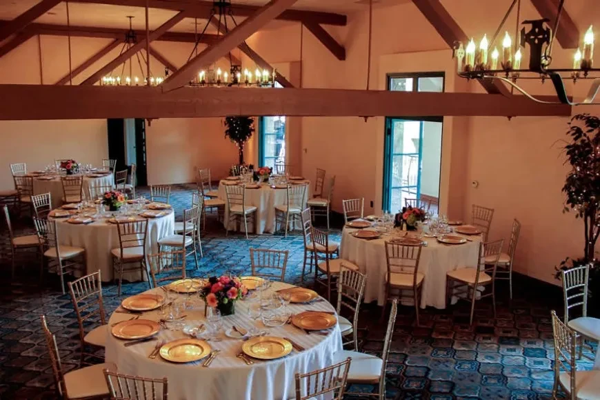 Elegant, empty event room with set tables, chandeliers, and wooden beams.