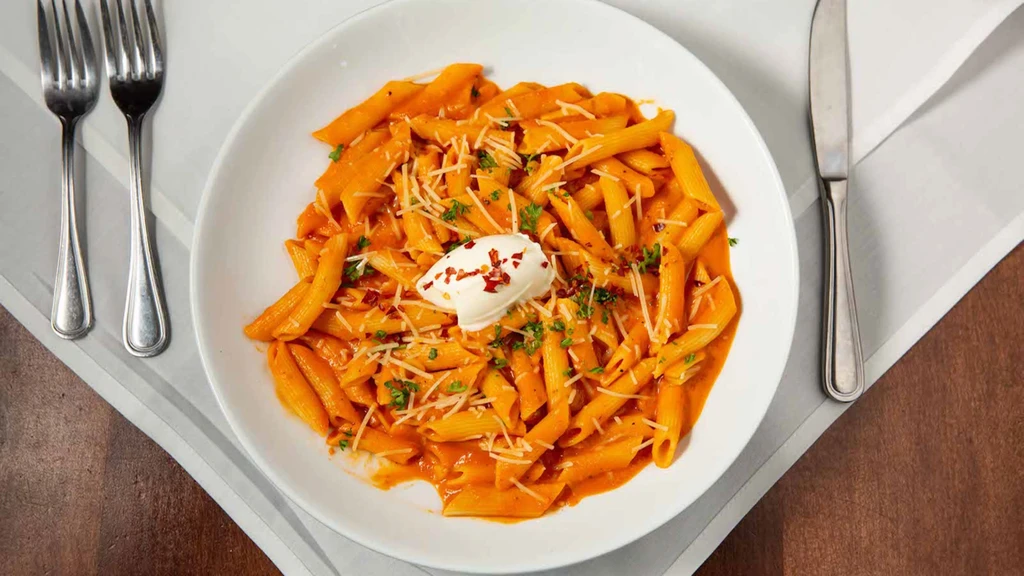 Penne pasta with creamy reddish-orange sauce, cheese, herbs, cutlery.