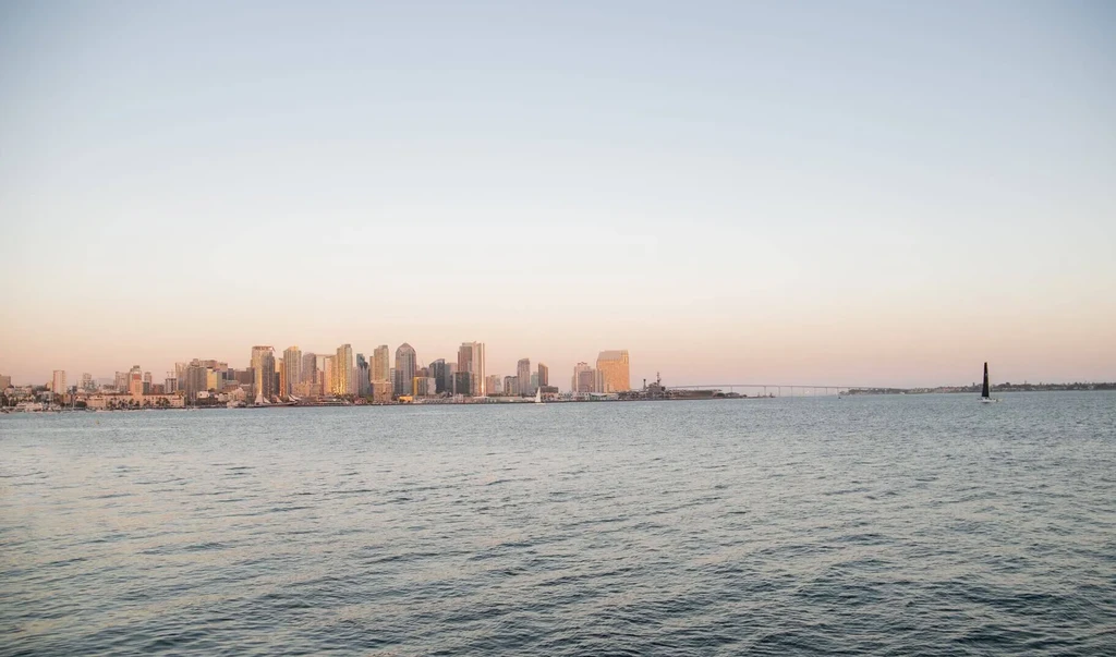 City skyline across water at sunset, with sailboat and bridge.