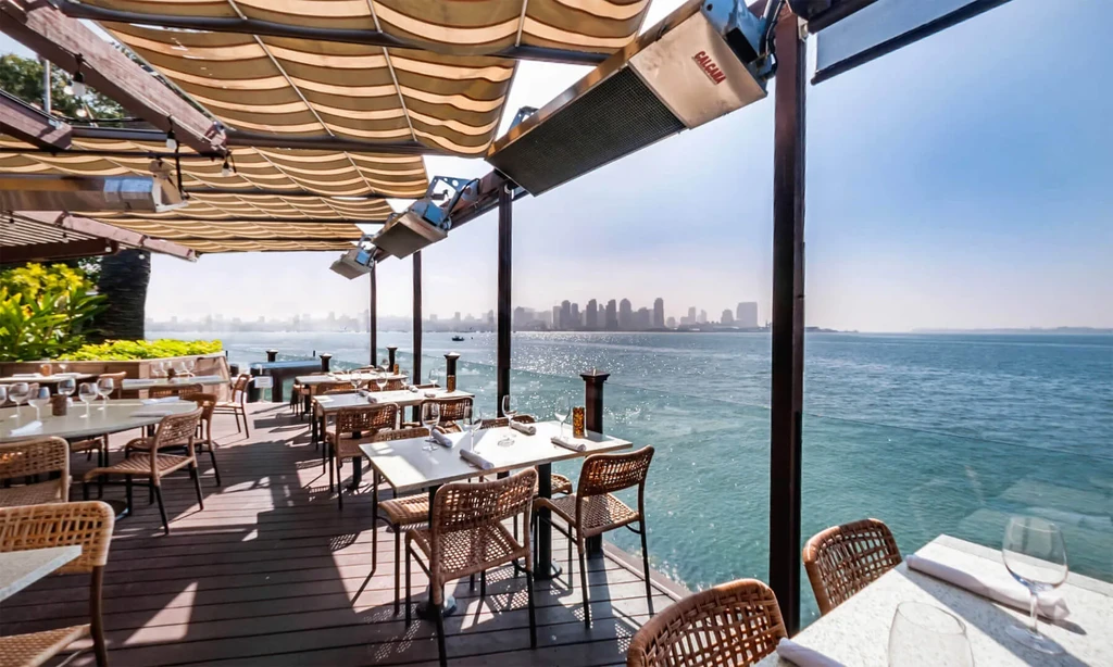 Outdoor restaurant deck overlooking a sunny bay and city skyline.