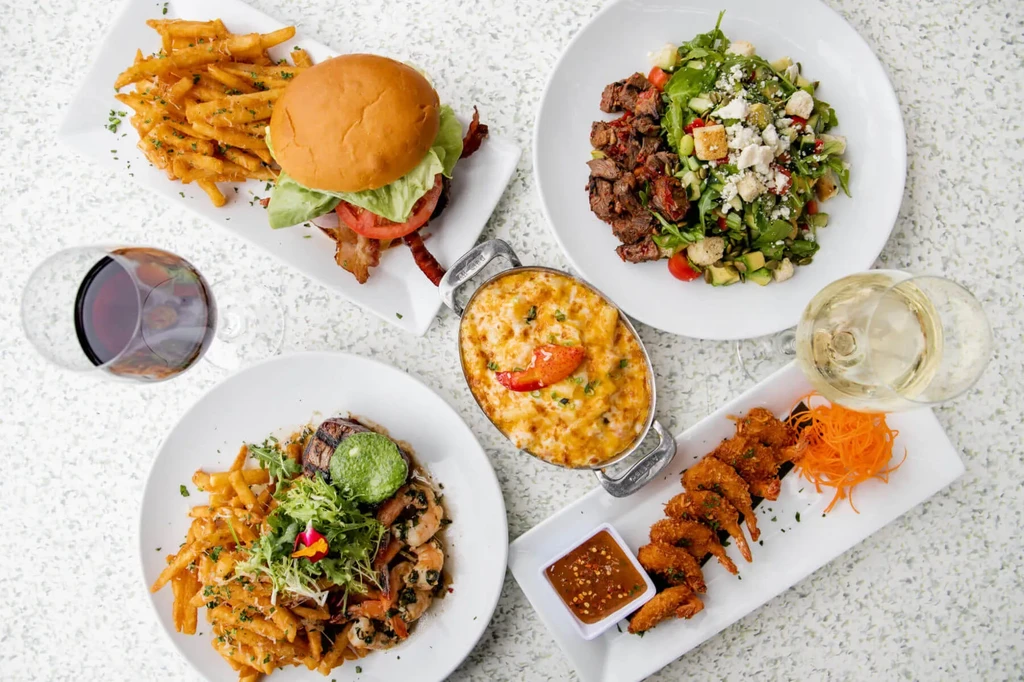 Table laden with diverse dishes: burger, salad, mac and cheese, shrimp, wine.