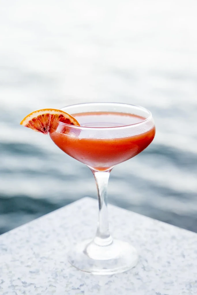 Reddish-orange cocktail in coupe, blood orange garnish, blurred water background.