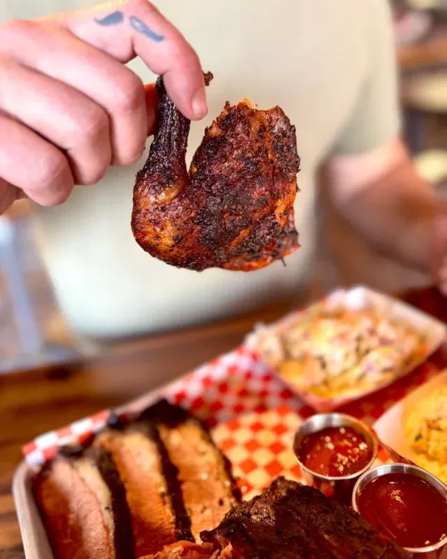 Hand with mustache tattoo holding a bitten, dark barbecued chicken.