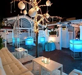 Outdoor lounge at night, lit tree, glowing blue tables, and white seating.