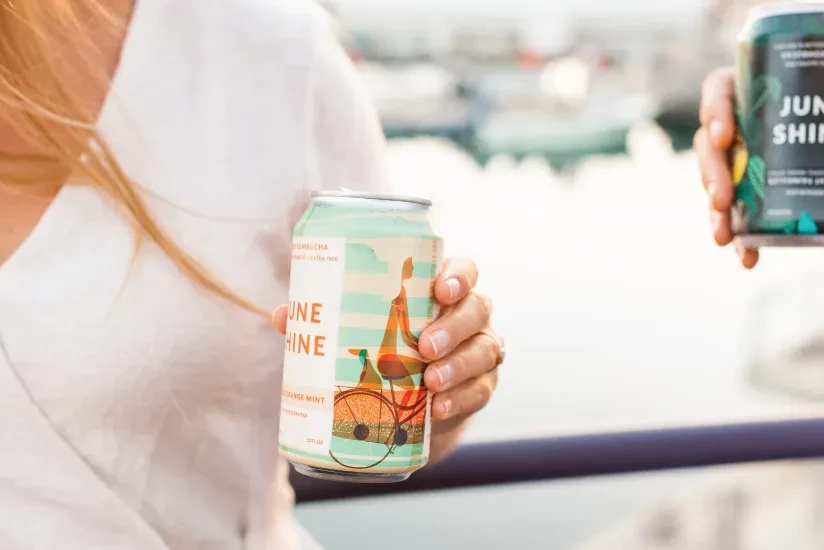 Hand holds JuneShine Orange Mint kombucha can outdoors; second can blurred.