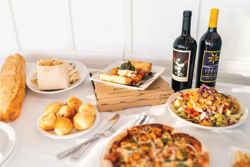 Spread of takeout food: pizza, burrata, salad, fries, and wine.
