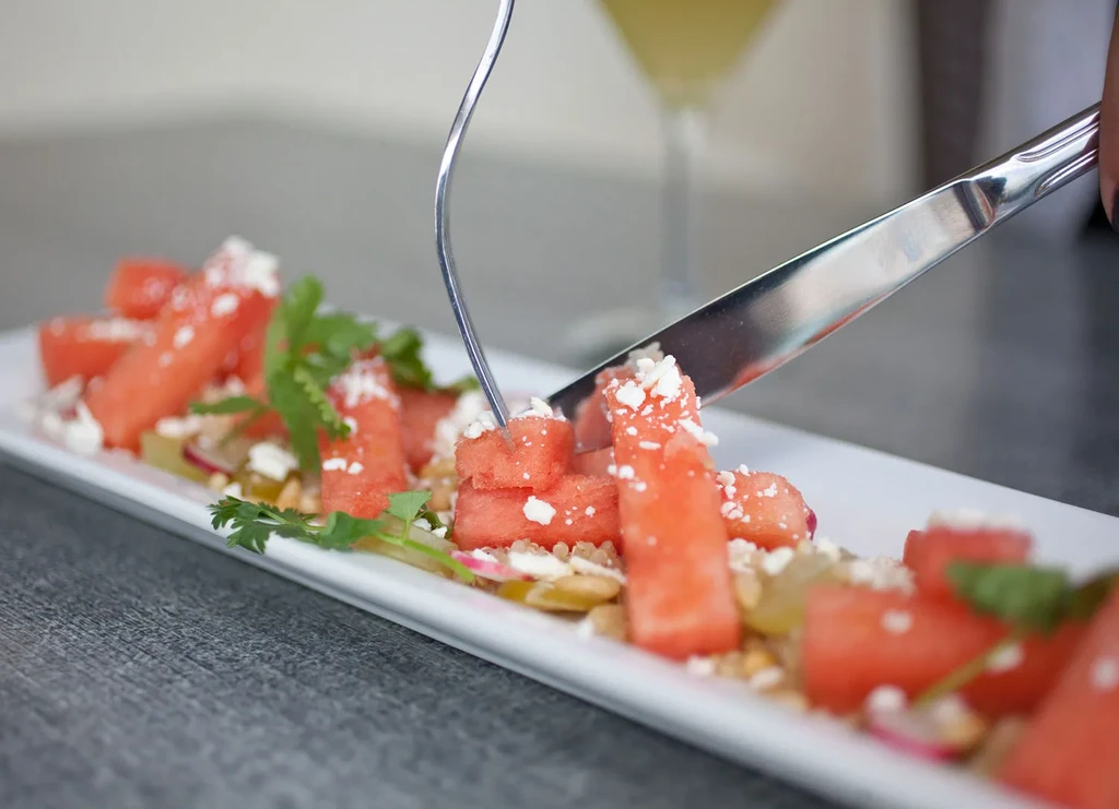 Plated watermelon salad with feta, herbs, and pine nuts; fork and knife.
