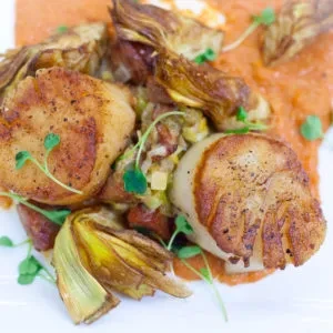 Golden pan-seared scallops, artichoke hearts, vegetables, microgreens on orange sauce.
