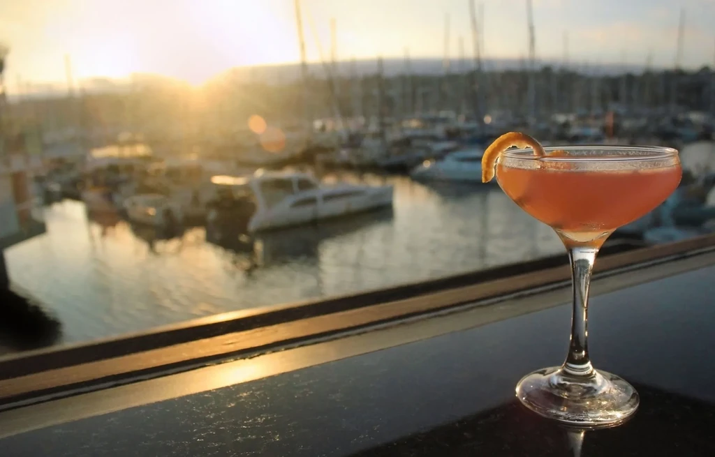 Orange cocktail in coupe glass, blurred marina sunset background.