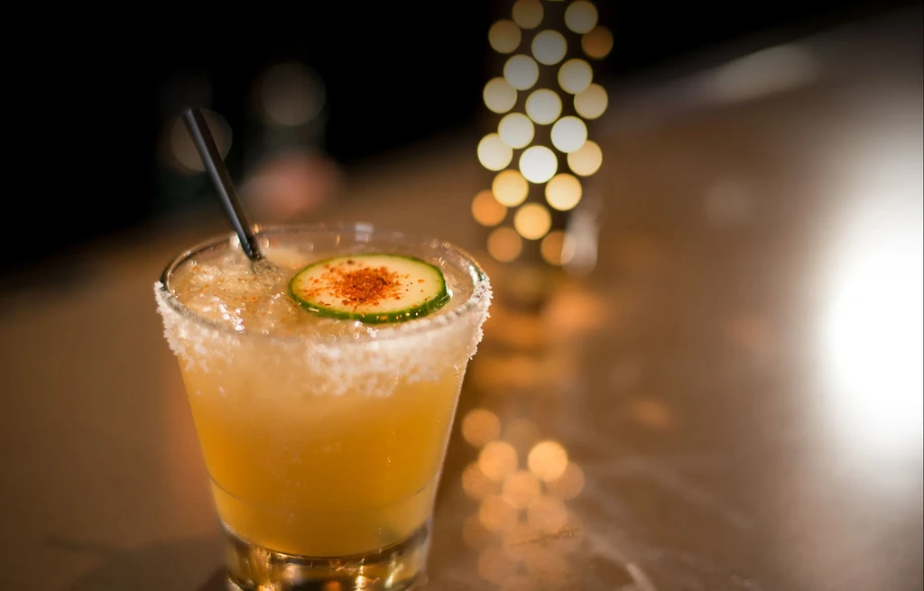 Close-up: Yellowish-orange cocktail, crushed ice, cucumber slice, spiced rim.