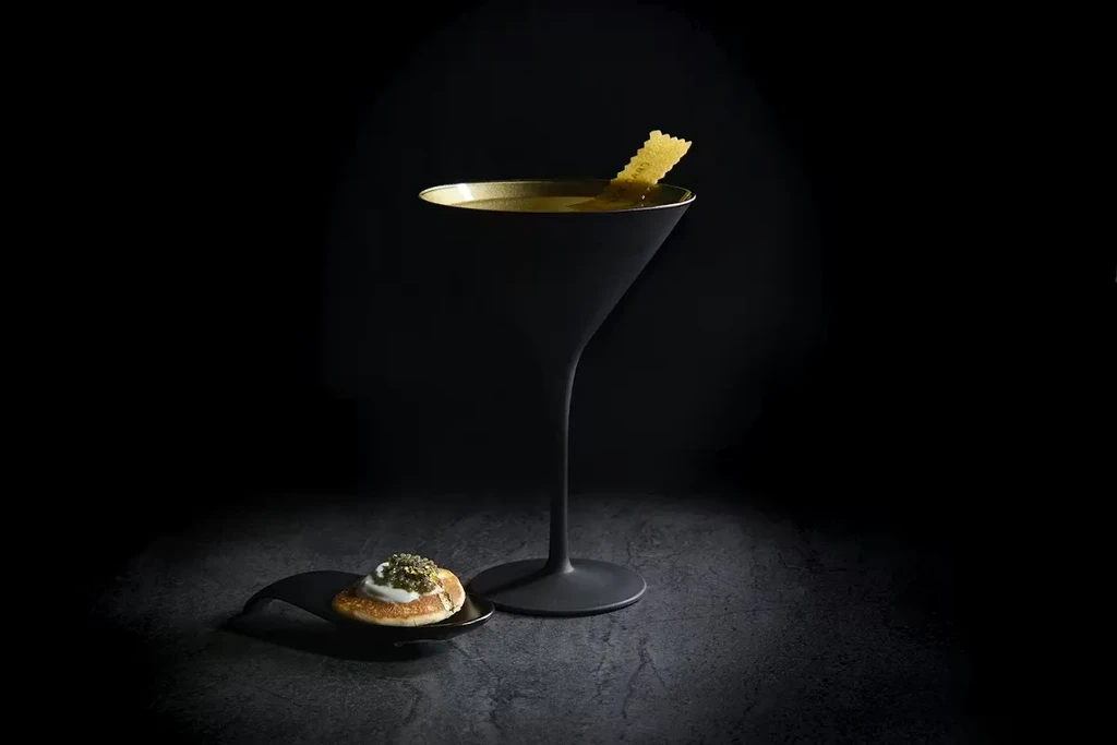 Martini in black and gold glass with crisp, blini with caviar.
