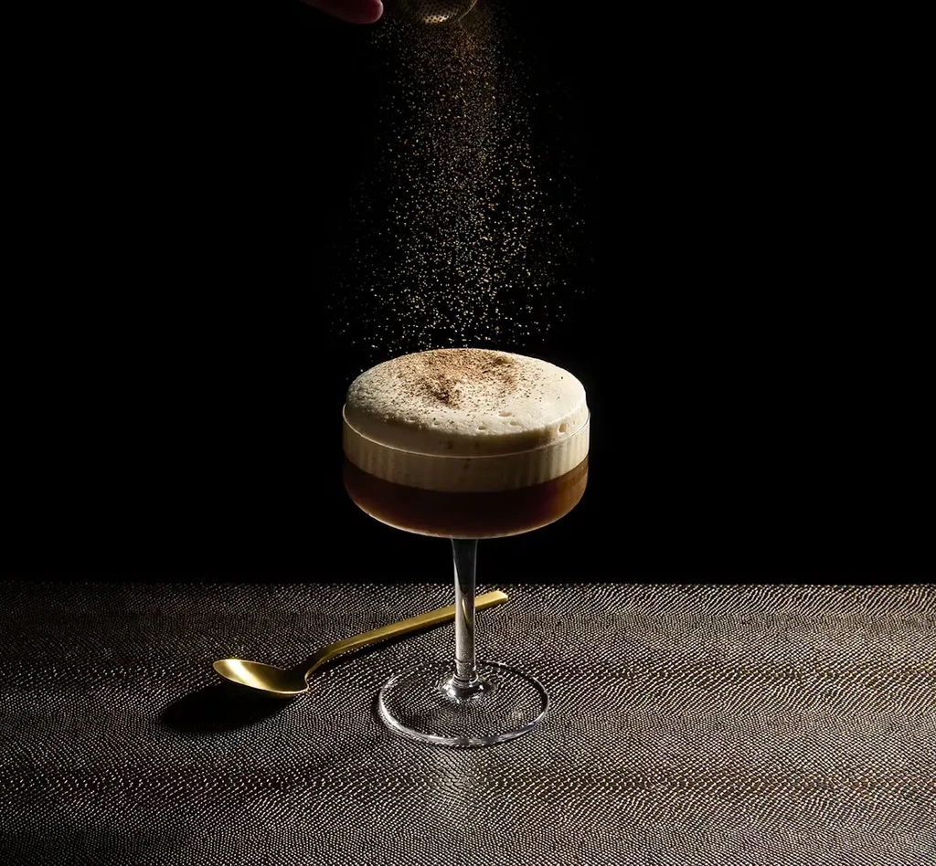 Dark layered drink in stemmed glass, brown powder sprinkled on foam.