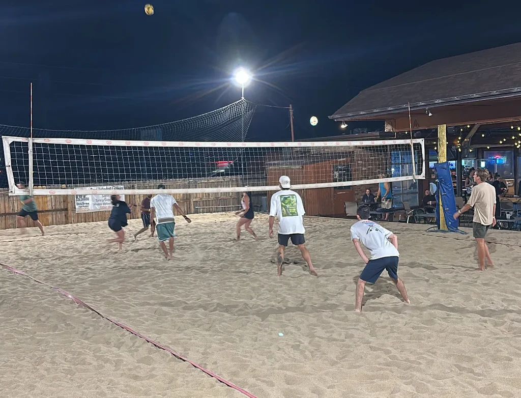 People playing night beach volleyball on a lit sand court and bar.