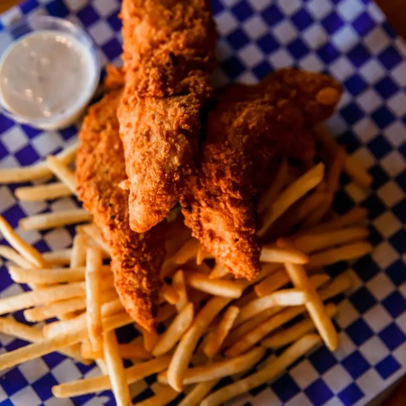 Three golden-brown fried chicken tenders, fries, and sauce on checkered paper.