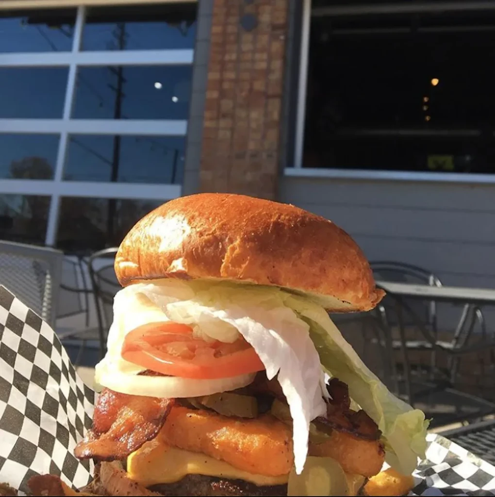 Close-up of a stacked burger with bacon, tater tots, and cheese outdoors.