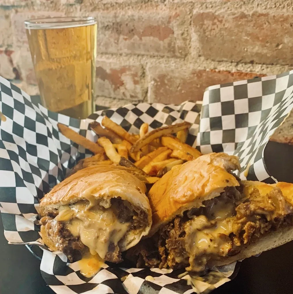 Overflowing cheesesteak, french fries, and beer in a basket.