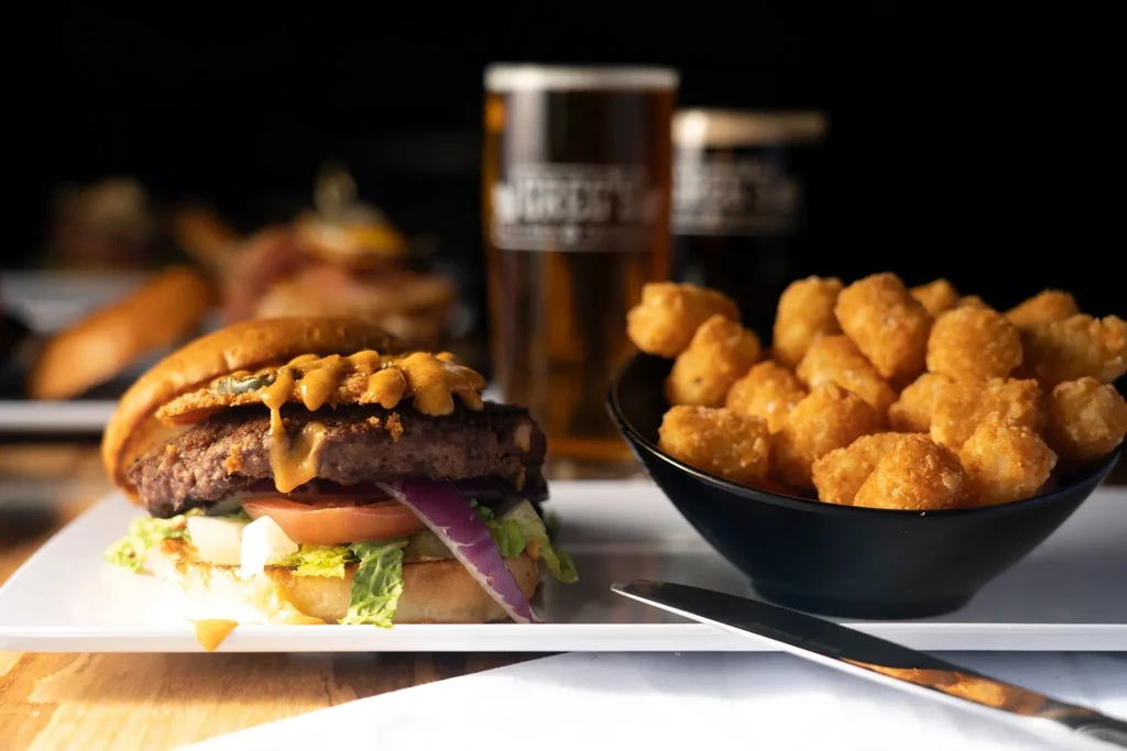 Open-faced burger, tater tots, and two beers in a dimly lit pub.