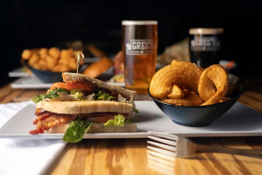Club sandwich halves, curly fries, and two GREG'S beers on a table.