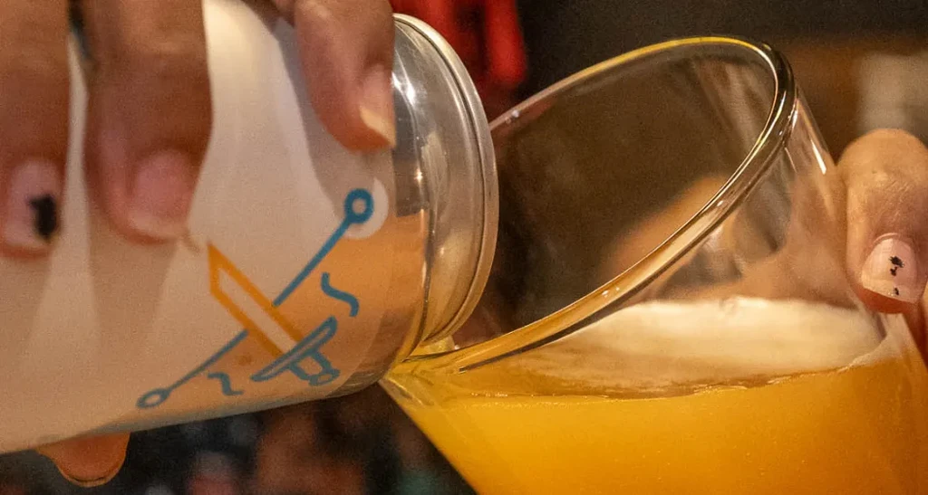 Person's hands pour hazy yellow drink from can into glass.