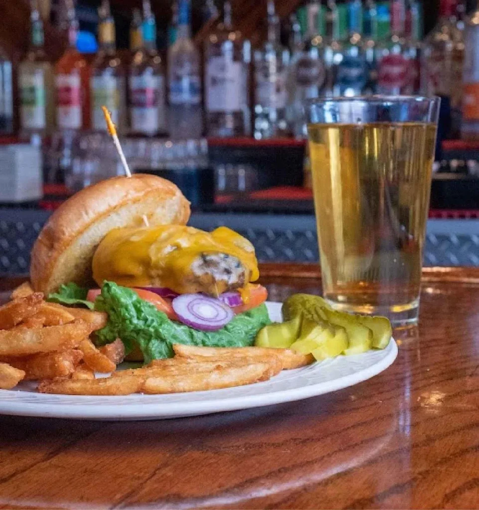 Partially deconstructed burger, fries, pickles, and beer at a bar.