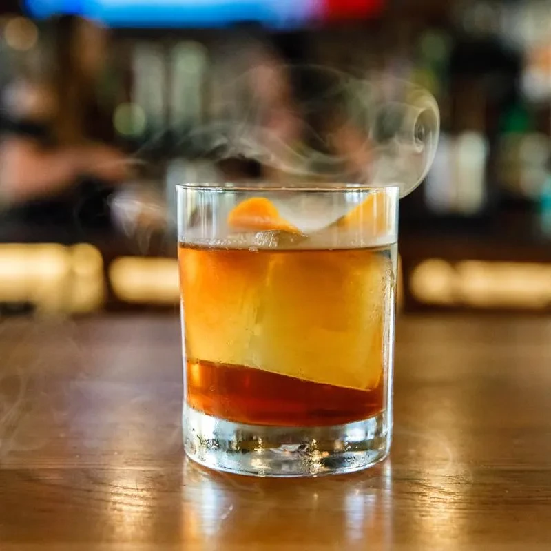Amber cocktail with orange peel and smoke rising from a rocks glass.