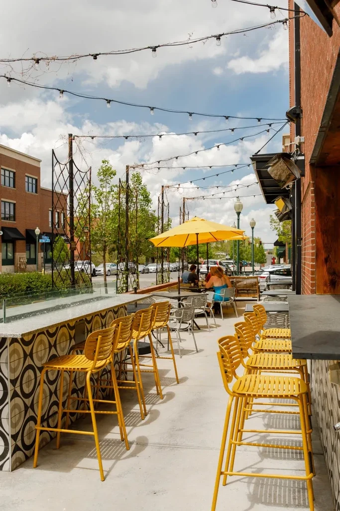 Outdoor restaurant patio with yellow bar stools, umbrella, people, and string lights.