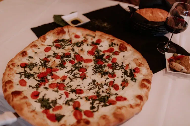 Close-up overhead pizza with tomatoes, basil, wine, and appetizers.