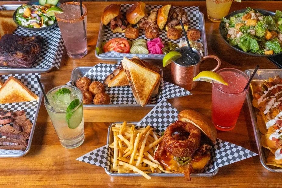 Overhead spread of roasted meats, nachos, burgers, salads, and drinks.