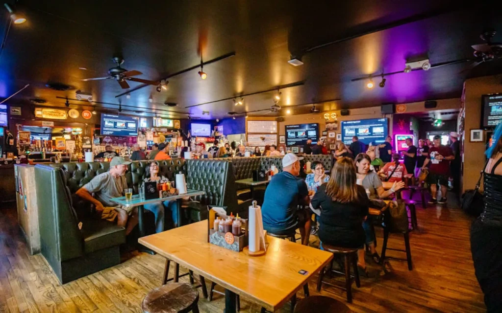 Lively bar interior with dark green booths, wooden tables, many patrons.