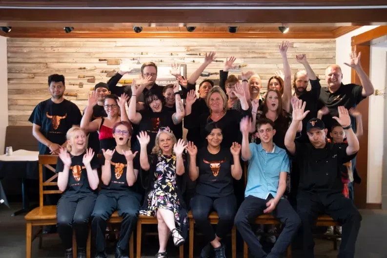 Smiling group of twenty-plus in bull skull shirts wave in a restaurant.