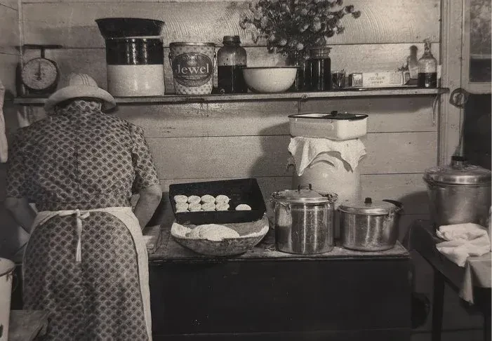 Person in apron bakes biscuits in a busy vintage kitchen.